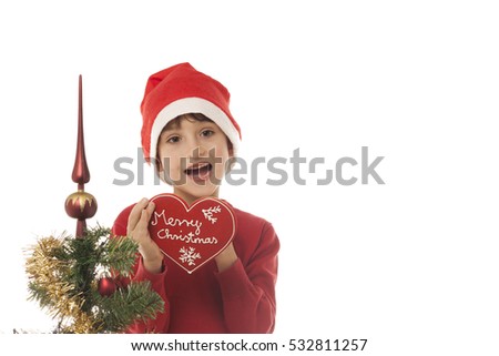 Schoolboy dressed in red with a santa hat isolated on white background. Christmas tree in the background. Child holding a red heart gingerbread with Merry Christmas written. Space left for copy