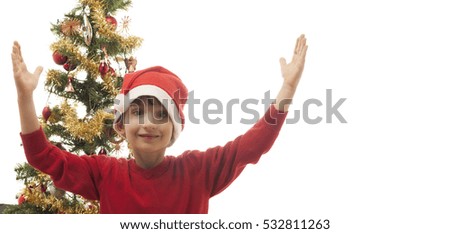Schoolboy dressed in red with a santa hat isolated on white background. Christmas tree in the background. Child smiling with hands in the air. Space left for copy