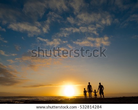 Family at sunset by the ocean. People hold hands and look at the setting sun. We see people from the back.