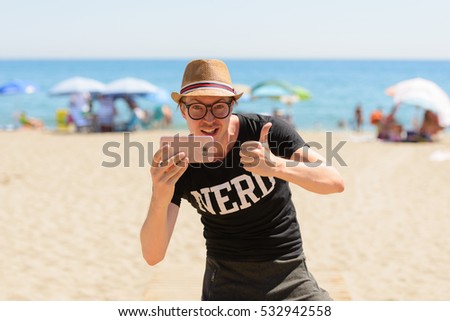 Young handsome Caucasian nerd taking picture and giving thumb up on vacation at beach in Spain