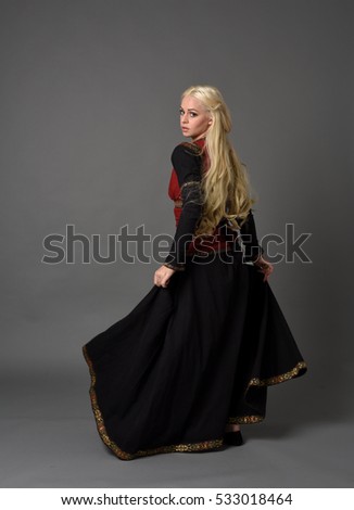 full length portrait of a beautiful blonde woman wearing a black and red medieval gown, standing pose against a grey studio background. Royalty-Free Stock Photo #533018464