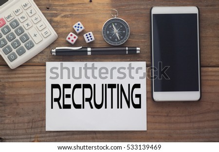 Recruiting written on paper,Wooden background desk with calculator,dice,compass,smart phone and pen.Top view conceptual.