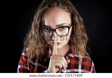 Portrait of teen girl with curly blond hair, wearing glasses and red plaid shirt making silence sign- finger on lips for silence. Human emotion facial expression feelings, body language reaction.