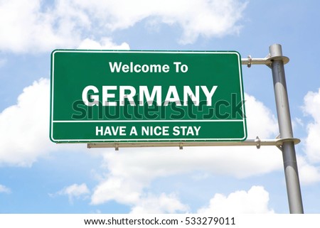 Green overhead road sign with a Welcome to Germany, Have a Nice Stay concept against a partly cloudy sky background.