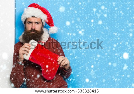 young handsome bearded santa claus man with long beard in red sweater and new year hat holds decorative christmas or xmas stocking on blue background under white snow and snowflakes, copy space