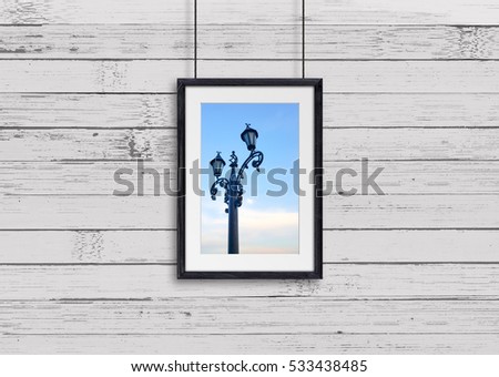 Black wooden frame, hanging on cords against old painted panels wall, with city motif picture, lamp post on blue sky background. Gallery style design, street cafe decor mock up