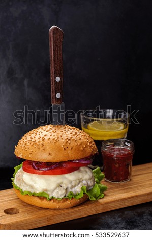 Fresh homemade burger with chicken burger cutlet, tomato sauce and mozzarella cheese in traditional buns, served on wood chopping board with vintage knife over dark background