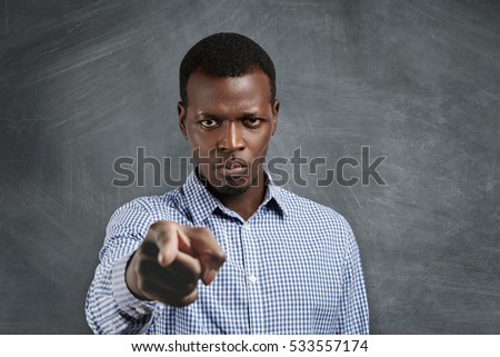 Portrait of unhappy African boss having mad expression pointing his index finger at camera, looking angrily and frowning as if accusing or blaming you for mistake. Selective focus on man's face Royalty-Free Stock Photo #533557174