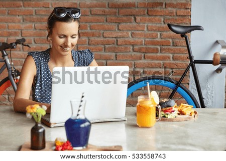 Attractive woman photographer retouching pictures using photo editor, having lunch, sitting in front of generic laptop. Student girl studying online on notebook, smiling joyfully while making progress