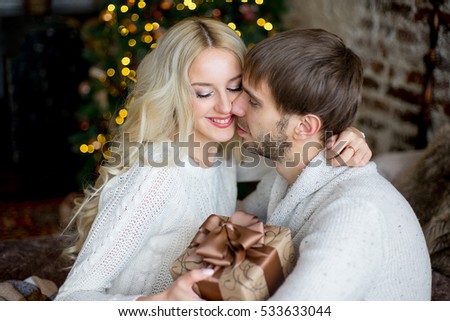 Happy couple of lovers in white pullovers give each other gifts sitting on the bed. Christmas tree and new year gift at home. Young family together