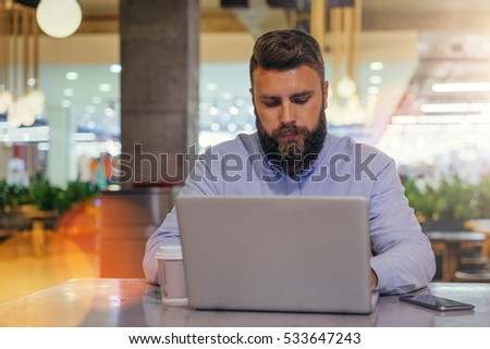 Front view.Young bearded businessman wearing blue shirt,sitting at table in cafe and uses laptop.On desk smartphone and cup of coffee.Man browsing internet on computer.Freelancer working.Film effect