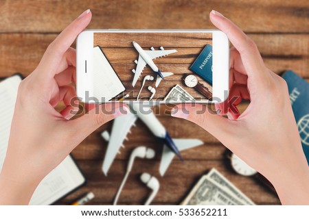 Woman hand using smart phone taking photo for preparation traveling with pencil, watch, money, passport, airplane, noted book and  earphone on vintage wooden background. Travel concepts.