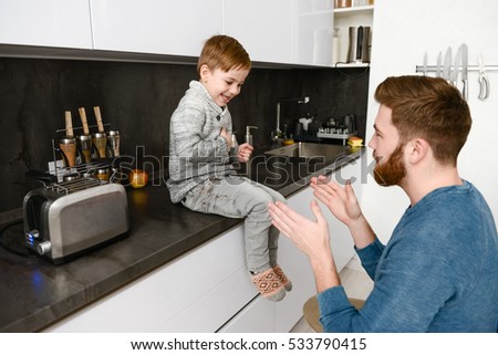 Picture of cheerful father dressed in blue sweater playing with his little cute son in kitchen
