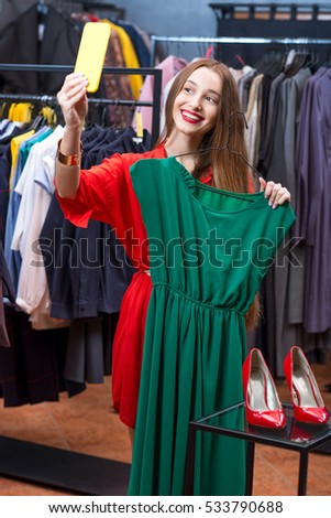 Young woman making selfie photo with mobile photo trying green dress in the luxury clothing store