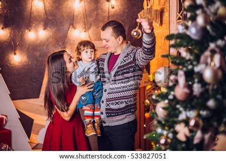 Modern Christmas Family Portrait sitting and smiling In Home Holiday Living Room, Mother, father and baby girl .Present Gift Box, House Decorating By lights Xmas Tree Candles Garland. Box with gifts.