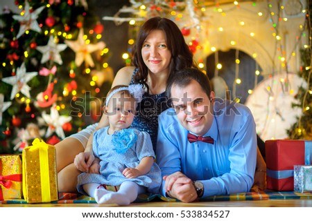 Happy family, sit on a floor near a fir-tree smile and pose on the camera. On a background a set of bright fires. Waiting for a holiday.