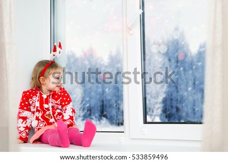 Cute little girl sitting by the window and looking at the winter forest. 