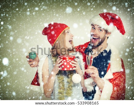 Cheerful young couple of pretty cute girl or woman and handsome bearded man in santa suits with new year tinsel bauble gift and glass of wine on white background under snow and snowflakes
