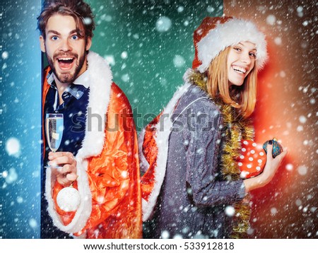 happy couple of pretty cute girl or woman and handsome bearded man in santa claus coat and hat with new year tinsel and gift box wine glass and xmas decoration on colorful backdrop under snow