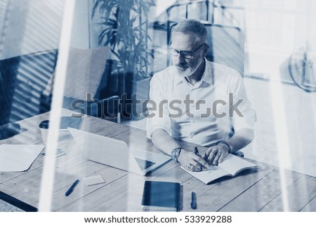 Concept of adult successful businessman wearing a classic glasses and working at the wood table in modern coworking place.Double exposure,skyscraper office building blurred background