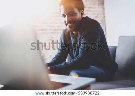 Cheerful bearded African man working on laptop while sitting sofa at his modern office place.Concept of young people using mobile devices.Brick wall blurred background.Horizontal, flares Royalty-Free Stock Photo #533930722