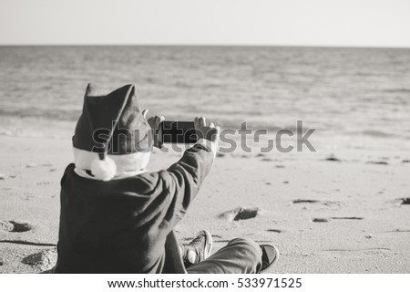 Back Side View of Joyful Child Santa Claus Using Smart Phone on Sunny Sky Ocean Beach Outdoors Background. Mock up screen, playing game on winter tropic holiday vacation. Black and white picture
