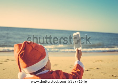 Back Side View of Joyful Child Santa Claus Using Smart Phone on Sunny Blue Sky Ocean Beach Outdoors Background. Mock up screen, taking photo, playing game on winter tropic holiday vacation