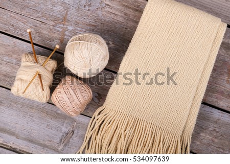 Beige knitted scarf with fringe on wooden background. Items for knitting and needlework.