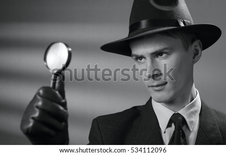 Handsome young vintage detective wearing a hat holding a magnifying glass in his gloved hand as he searches for clues, black and white portrait Royalty-Free Stock Photo #534112096