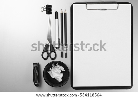 Black and white stationery on gray desk: clipboard with blank paper sheet, pencils, scissors, stapler, clip, pen, pen pot with paper trash. Top view point.