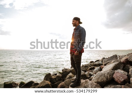 Picture of handsome african man wearing cap walking on the beach while listening music with earphones. Look at sea.