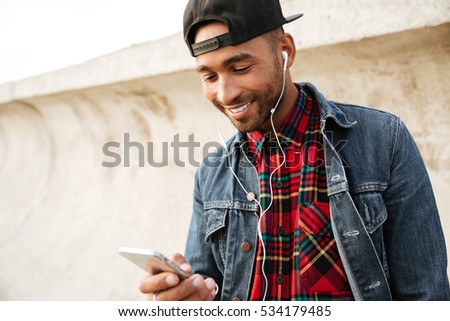 Picture of cheerful african young man wearing cap walking on the beach and chatting by his phone while listening music with earphones.