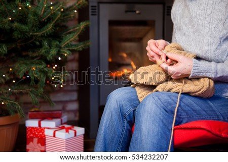 close up on old woman's hands sitting in red armchair and knitting - burning fireplace and Christmas tree with presents in the backgroung- cozy home concept