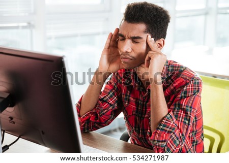 Picture of tired african guy with headache dressed in shirt in a cage print sitting in room while touching his head with hands.