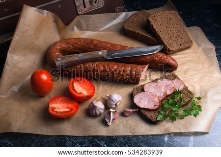 Close up view slices of smoked sausage with parsley and rye black bread, several tomatoes and garlic on the brown packaging paper. Vintage knife.
