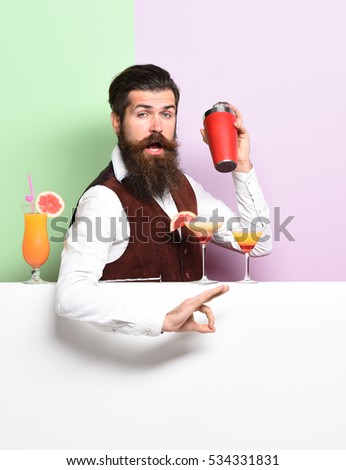 handsome bearded barman with long beard and mustache has stylish hair on funny face holding shaker and made alcoholic cocktail in vintage suede leather waistcoat on purple green studio background Royalty-Free Stock Photo #534331831