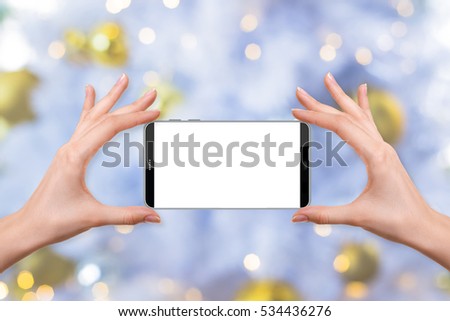 female holding a smartphone isolated blank screen with two hands on blur background of gold Christmas balls and decorations with warm light bokeh on a white christmas tree, ready for snap a picture