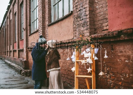 Young happy couple laughing and enjoying christmas street decorations with cookies, cacao, handmade wooden table, natural wreath and red brick wall