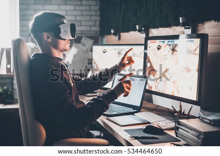 Testing games. Young man wearing virtual reality headset and gesturing while sitting at his desk in creative office Royalty-Free Stock Photo #534464650