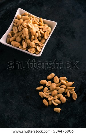 Vertical photo with top view on salted peanuts. Light brown nuts with salt placed on heap in front of white square bowl full of others. Nuts placed on dark black baking tray board with worn surface. 