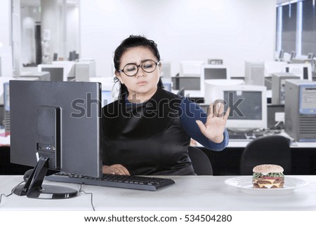 Picture of an overweight beautiful woman refusing burger on the plate and working with a computer in the office
