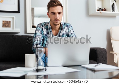 Picture of serious bristle man dressed in shirt in a cage print sitting on sofa in home and using laptop computer. Looking at laptop.