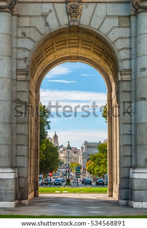 View of Metropolis building in Madrid, through an arch of the Plaza de la Independencia Monument.   Urban life, traffic flows in a busy, mid-day street scene. 
