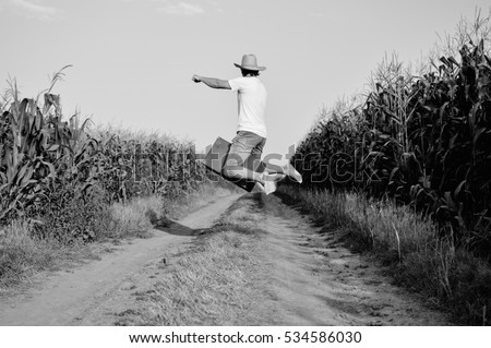 Black and white picture of joyful man jump with suitcase in country road field, back side view. Male looks like cowboy wearing straw hat and sitting on baggage, sunny countryside background.