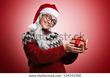 Smiling woman with long white hair wearing Santa hat, glasses and an ugly Christmas sweater looks longingly at a box with a present