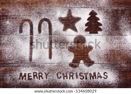 high-angle shot of a wooden table sprinkled with icing sugar or flour where you can read the text merry christmas and the silhouette of some candy canes a gingerbread man, a star and a christmas tree