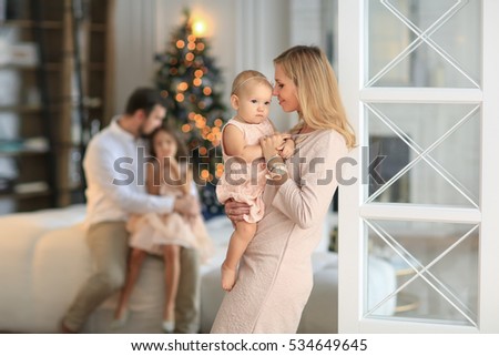 beautiful mother at the tender pink dress kissing little baby on hands sitting behind the father and daughter at the Christmas tree