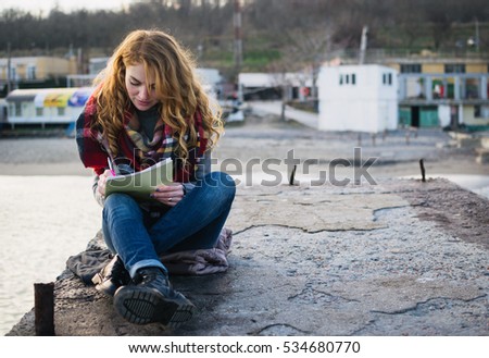 Young woman with red hair curls sitting and drawing with pen at the seacoast