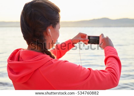 Active young woman listening to music during workout. Runner rests from routine exercise and makes photos. Happy jogger relaxing at the beach after run and looking at the sea.Back view