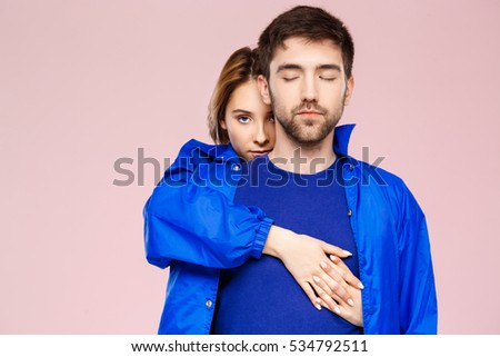 Funny young beautiful couple wearing one jacket embracing smiling over light pink background.